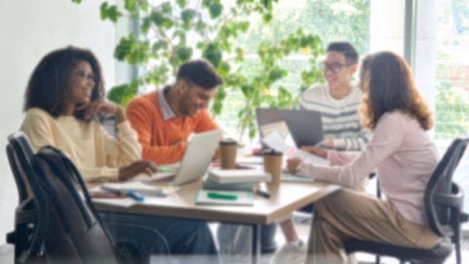 Four diverse young adults sitting around a table working on laptops and reviewing documents in a bright office with large windows and plants.