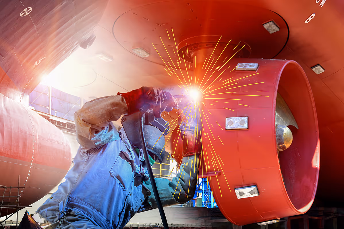 Worker welding a large red ship propeller in a dry dock.