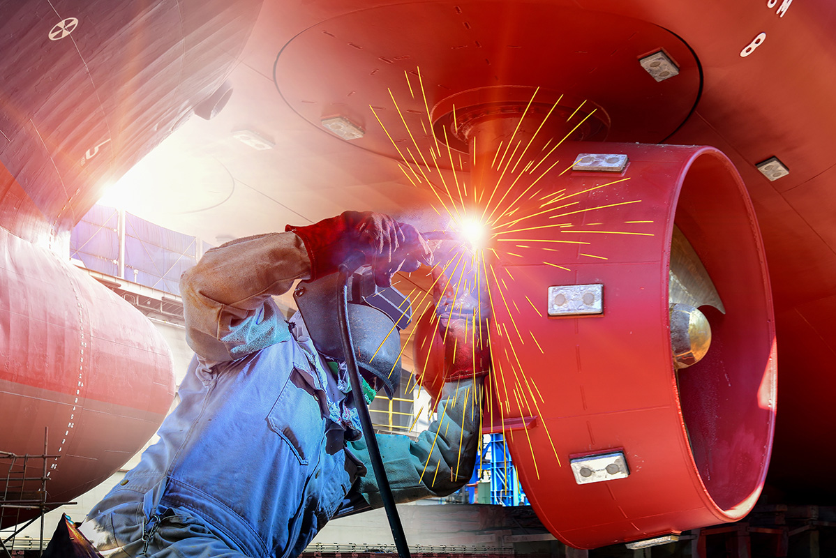 Worker welding a large red ship propeller in a dry dock.