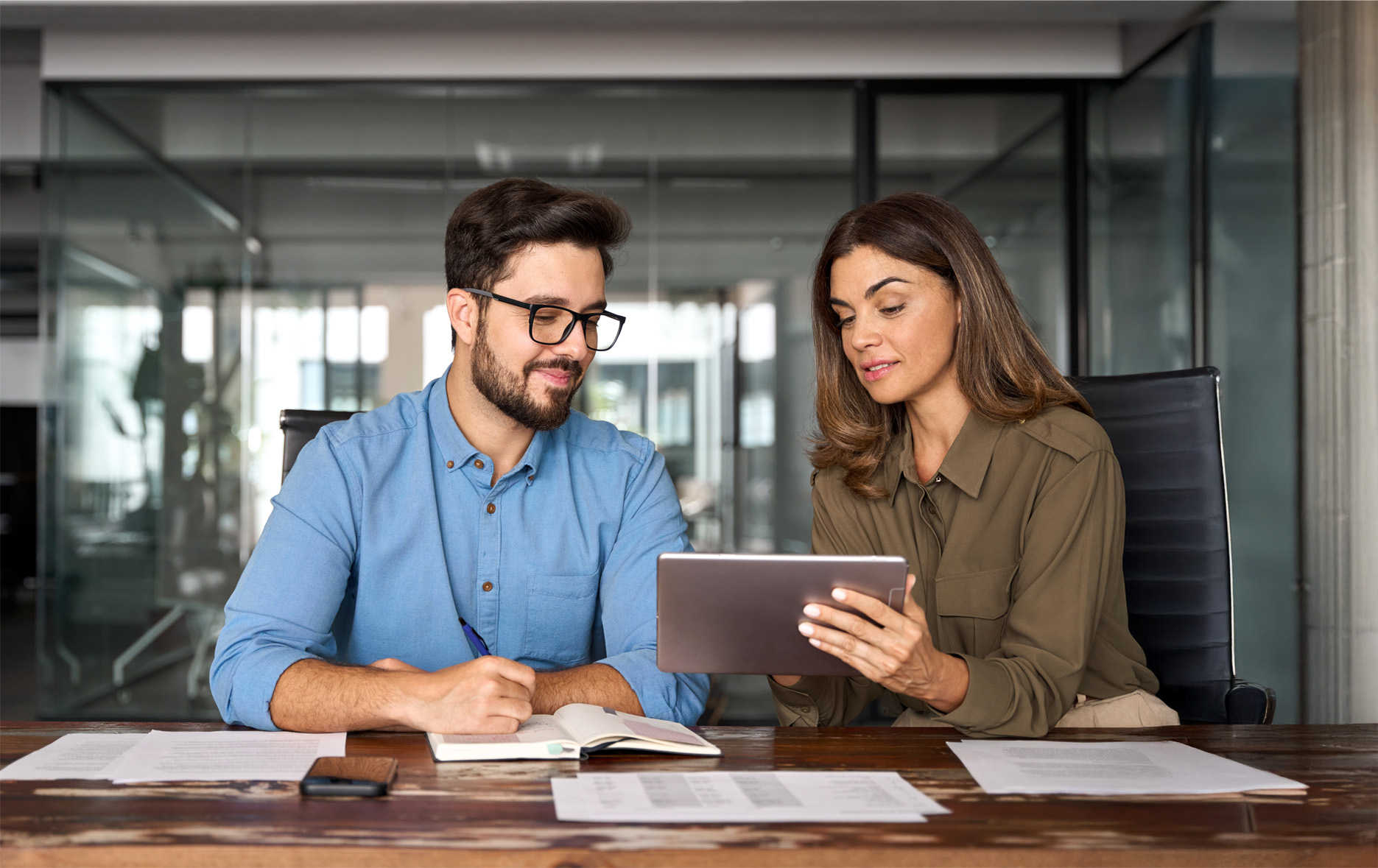 Man and woman sitting at a table in an office, the woman shows something on a tablet while the man takes notes in a notebook.