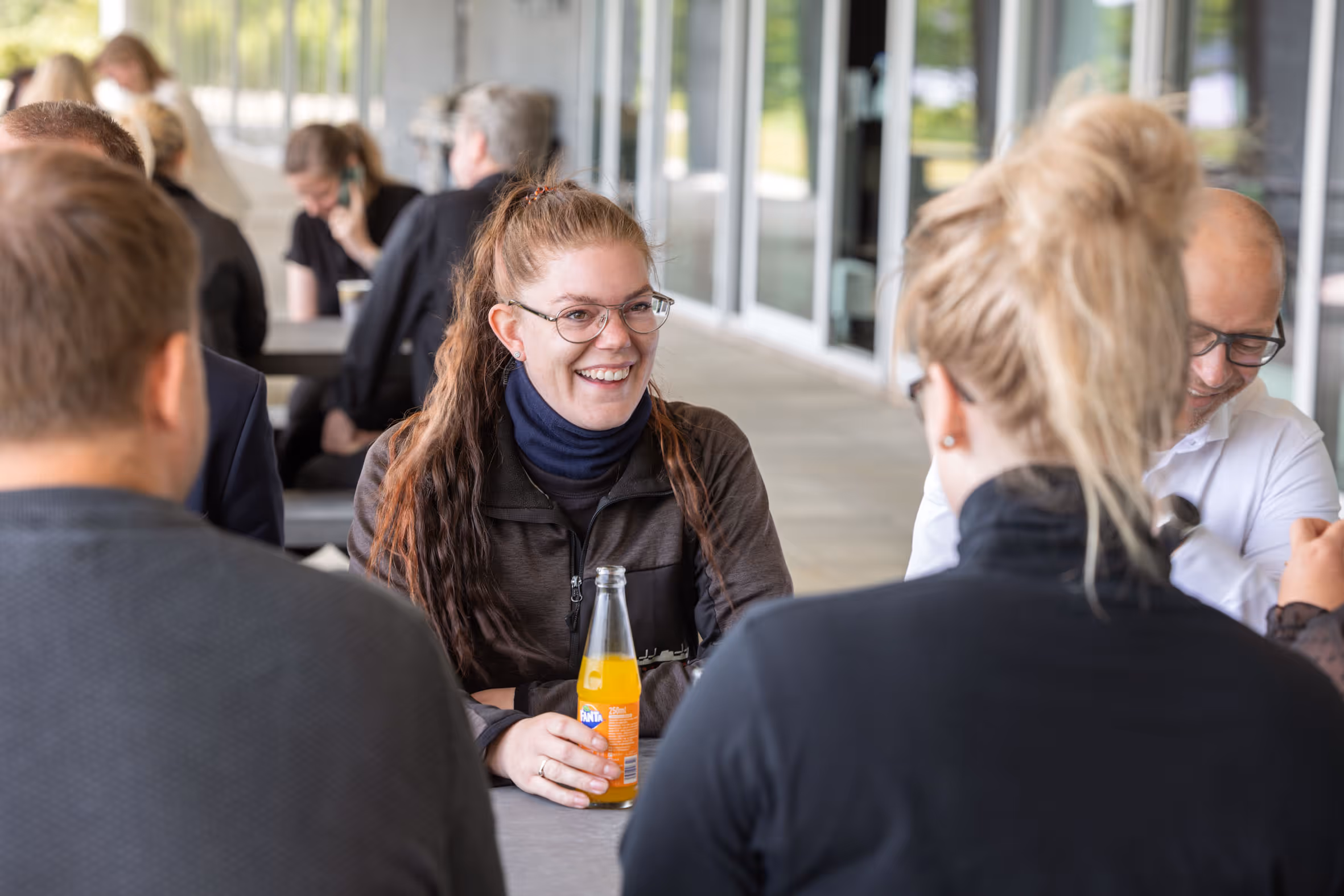 Group of Wrist employees sitting and chatting at an outdoor table, with a woman smiling and holding a bottle of orange Fanta soda.