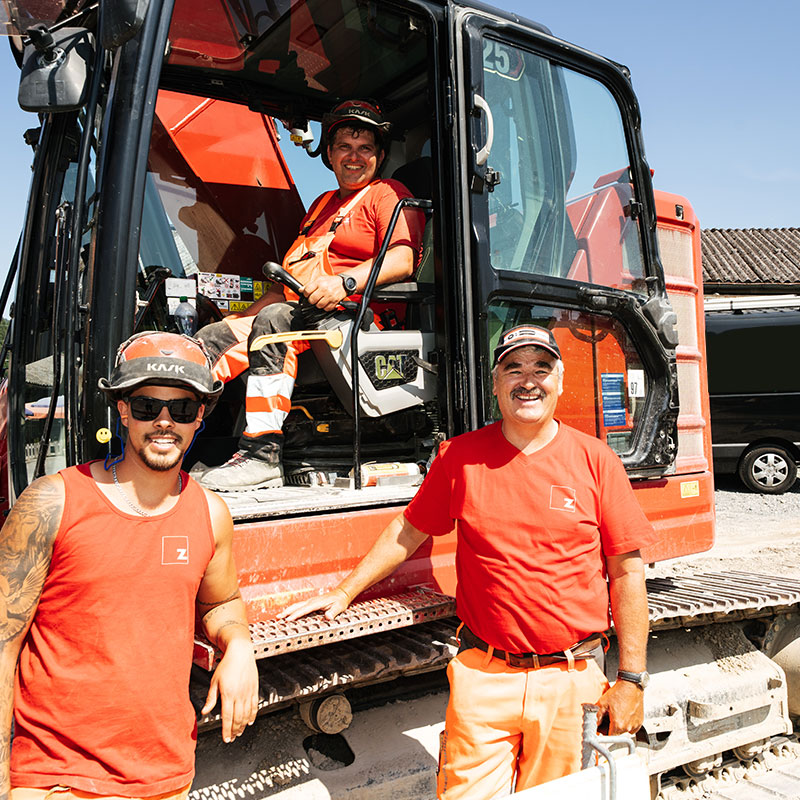 Zwei Mitarbeiter vor Bagger und einer im Bagger