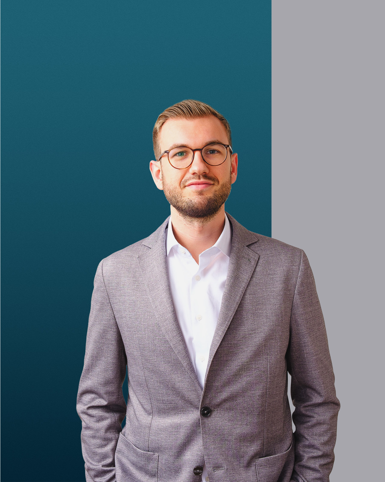Ein junger Mann mit Brille trägt grauen Anzug und weißes Hemd vor blau-grauem Hintergrund.