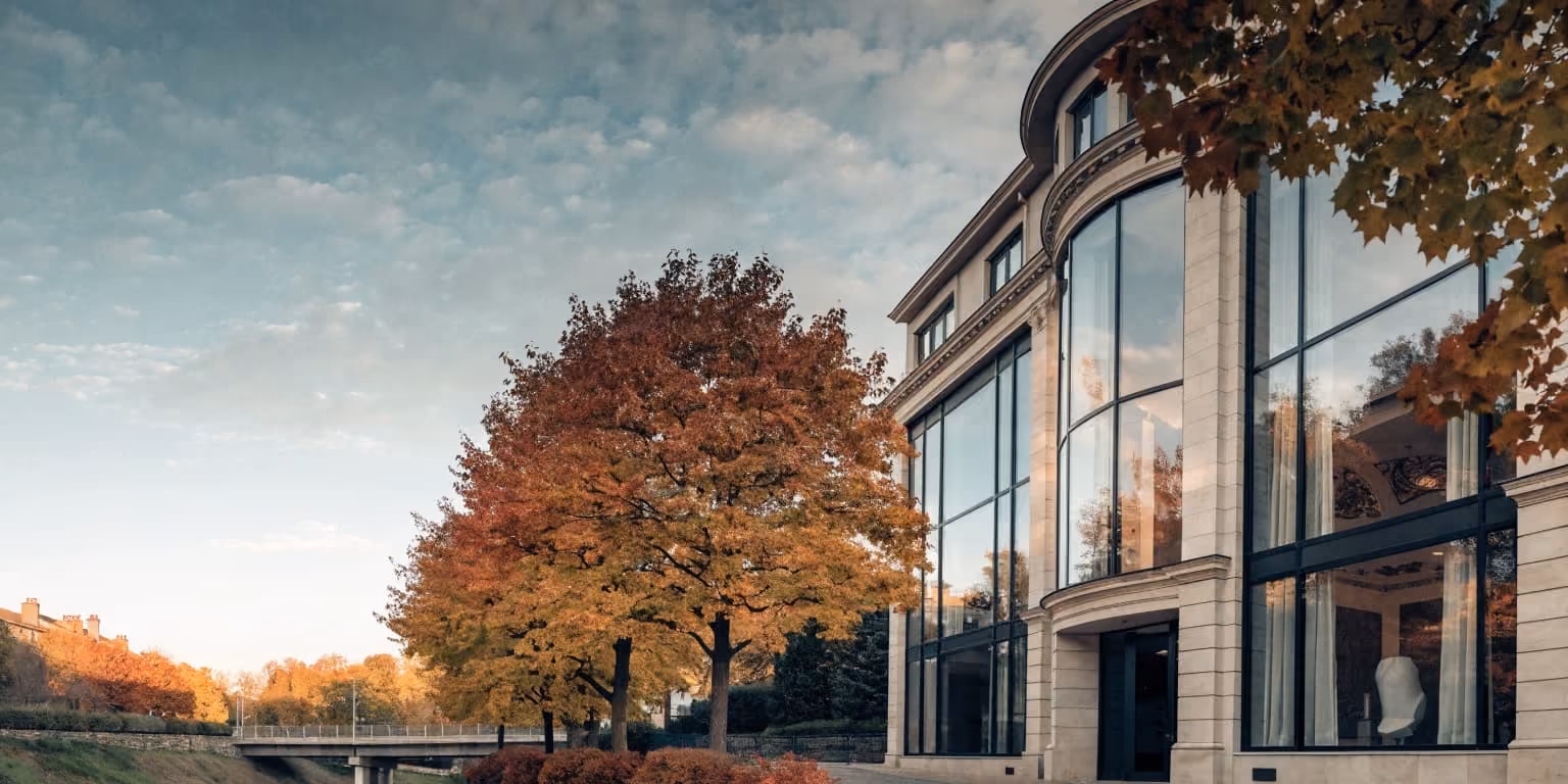 Modern building with large glass windows beside autumn trees with orange and yellow leaves under a cloudy sky.