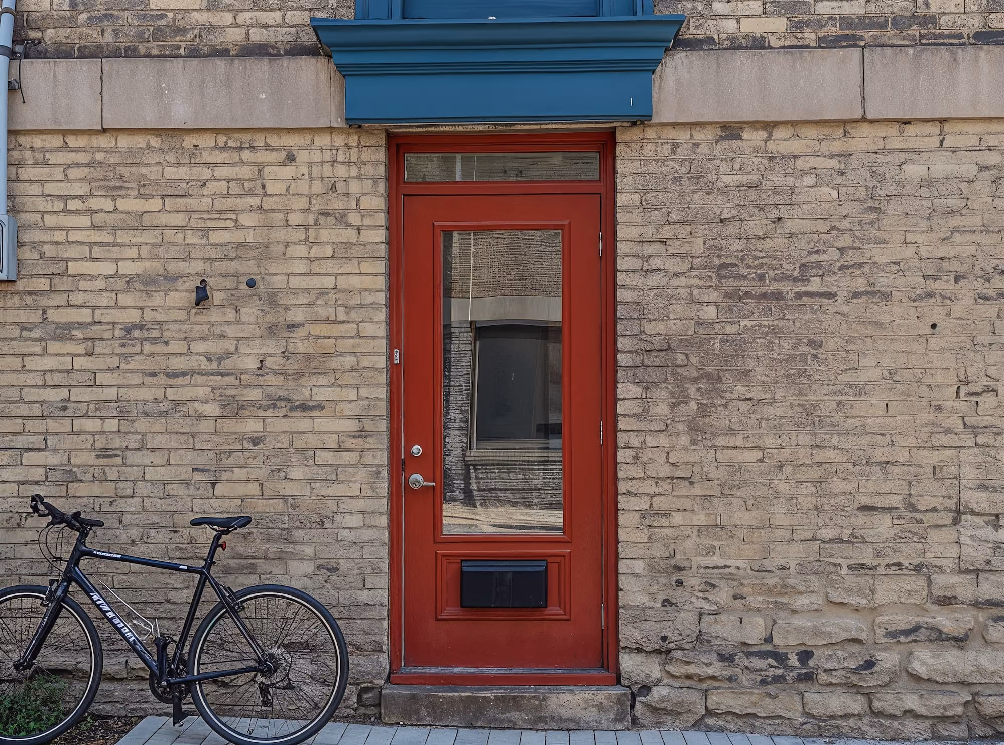 Black bicycle parked next to a red door set in a weathered brick wall with blue trim.