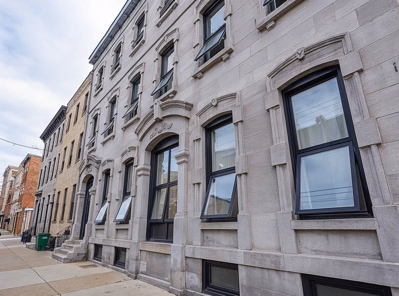 Row of old stone buildings with arched windows, some open, along a quiet urban sidewalk.