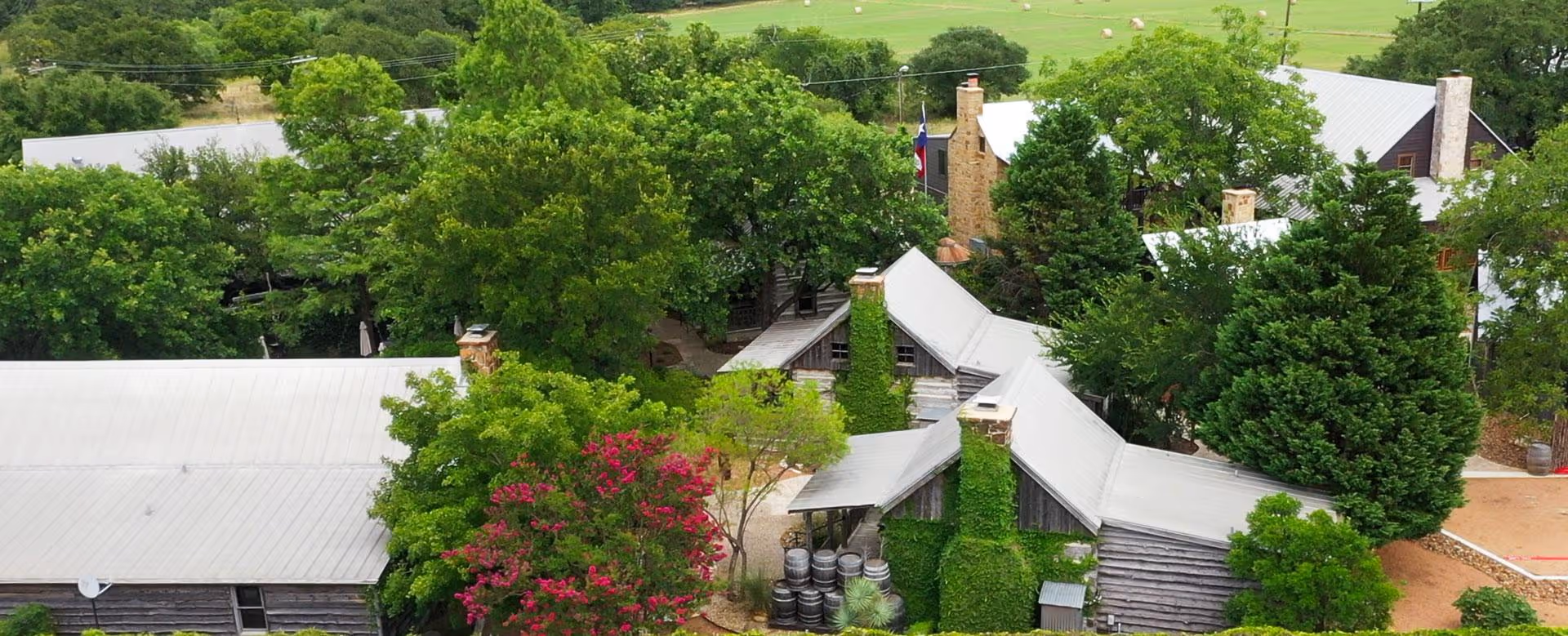 Aerial view of rustic wooden buildings surrounded by green trees and pink flowers