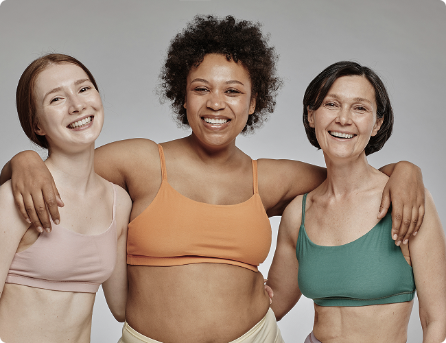 Three diverse women in colorful sports bras smiling and standing together, representing women’s health and body positivity.
