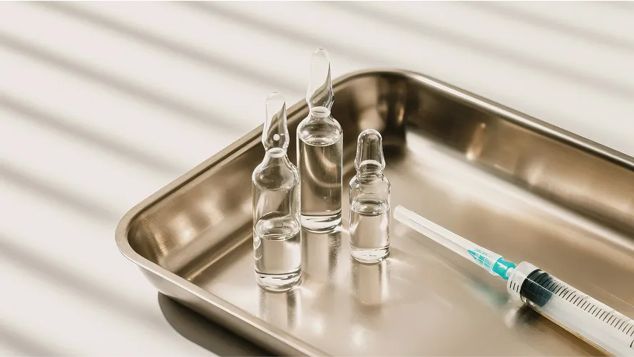 Three glass ampoules with clear liquid and a syringe on a metal tray, lit by soft shadowed light, representing long-acting injectable medications.