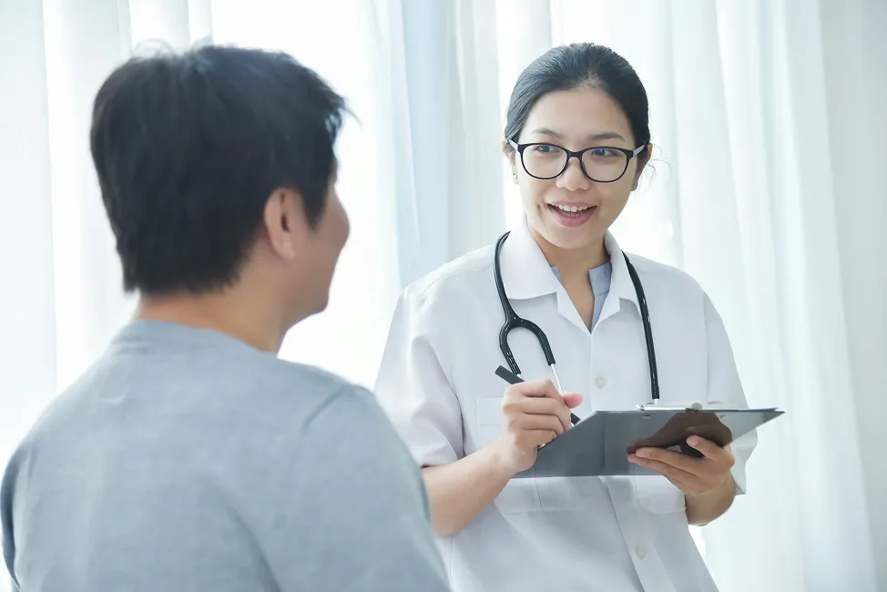 A SmartCareRxPharmacy healthcare provider wearing a white coat and stethoscope consults with a patient and writes on a clipboard in a brightly lit medical office.