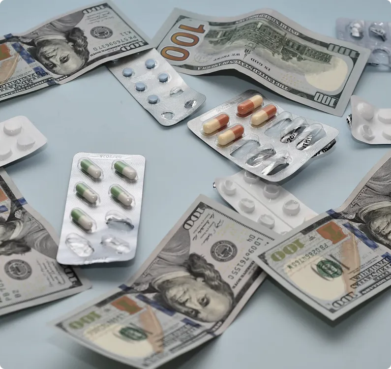  Various blister packs of pills and capsules are spread out among multiple one-hundred dollar bills on a light blue surface.