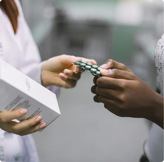 A pharmacist in a white coat hands a blister pack of green capsules to a patient, who reaches out to accept the medication while holding a white box, set in a pharmacy environment with a softly blurred background.