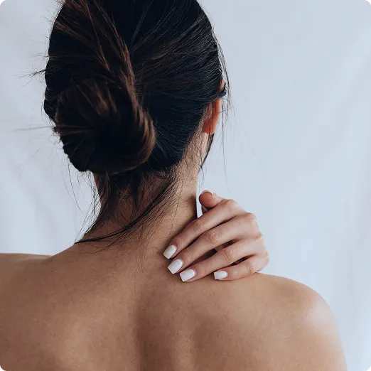 A woman with brown hair in a bun viewed from behind, her bare shoulder exposed, gently resting her hand with manicured nails on her neck and shoulder, evoking a sense of relief or self-care against a soft, neutral background.