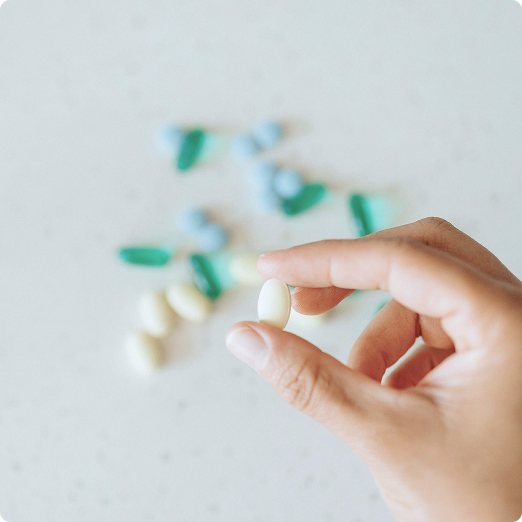 A close-up of a hand holding a single oval yellow pill between two fingers, with various blue, green, and yellow pills scattered in the background on a light speckled surface