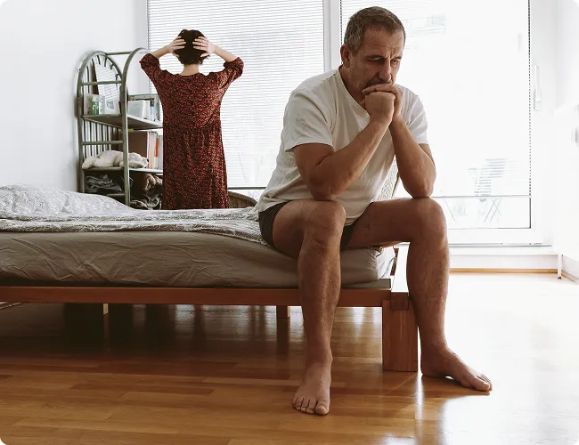 SmartCare Rx Pharmacy middle-aged man sitting at the edge of a bed appearing distressed, with a woman sitting on the other side, suggesting a couple facing intimacy or sexual health concerns