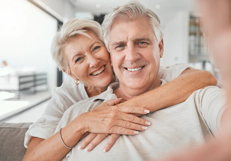 SmartCare Rx Pharmacy elderly couple sitting together on a couch in a warm, modern home, with the woman embracing the man from behind, highlighting companionship and support in senior health care