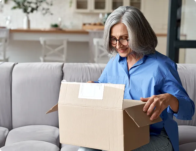 SmartCare Rx Pharmacy older woman with gray hair opening a cardboard package while sitting on a sofa in a modern, well-lit home, highlighting convenience and personalized pharmacy delivery service