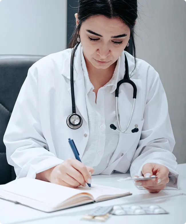 SmartCare Rx Pharmacy female doctor in a white coat writing notes at her desk with a stethoscope, emphasizing professional care.