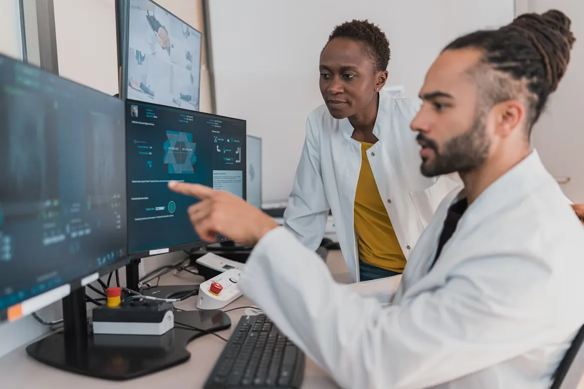 Two medical professionals in lab coats analyze diagnostic data and visuals on multiple computer monitors in a modern hospital control room, collaborating on patient care technology