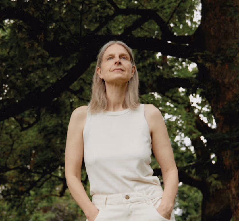 A woman with long gray hair, wearing a sleeveless white top and light pants, stands outdoors with her hands in her pockets. Lush green trees in the background highlight a serene scene of healthy aging and longevity.