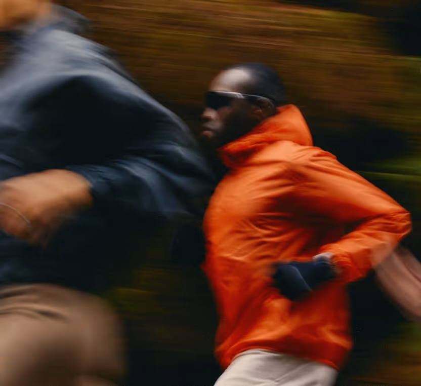 Two people are running outdoors, emphasizing fat burning and performance. The person in the foreground wears an orange jacket and sunglasses. Motion blur highlights their speed, with lush greenery visible in the background.