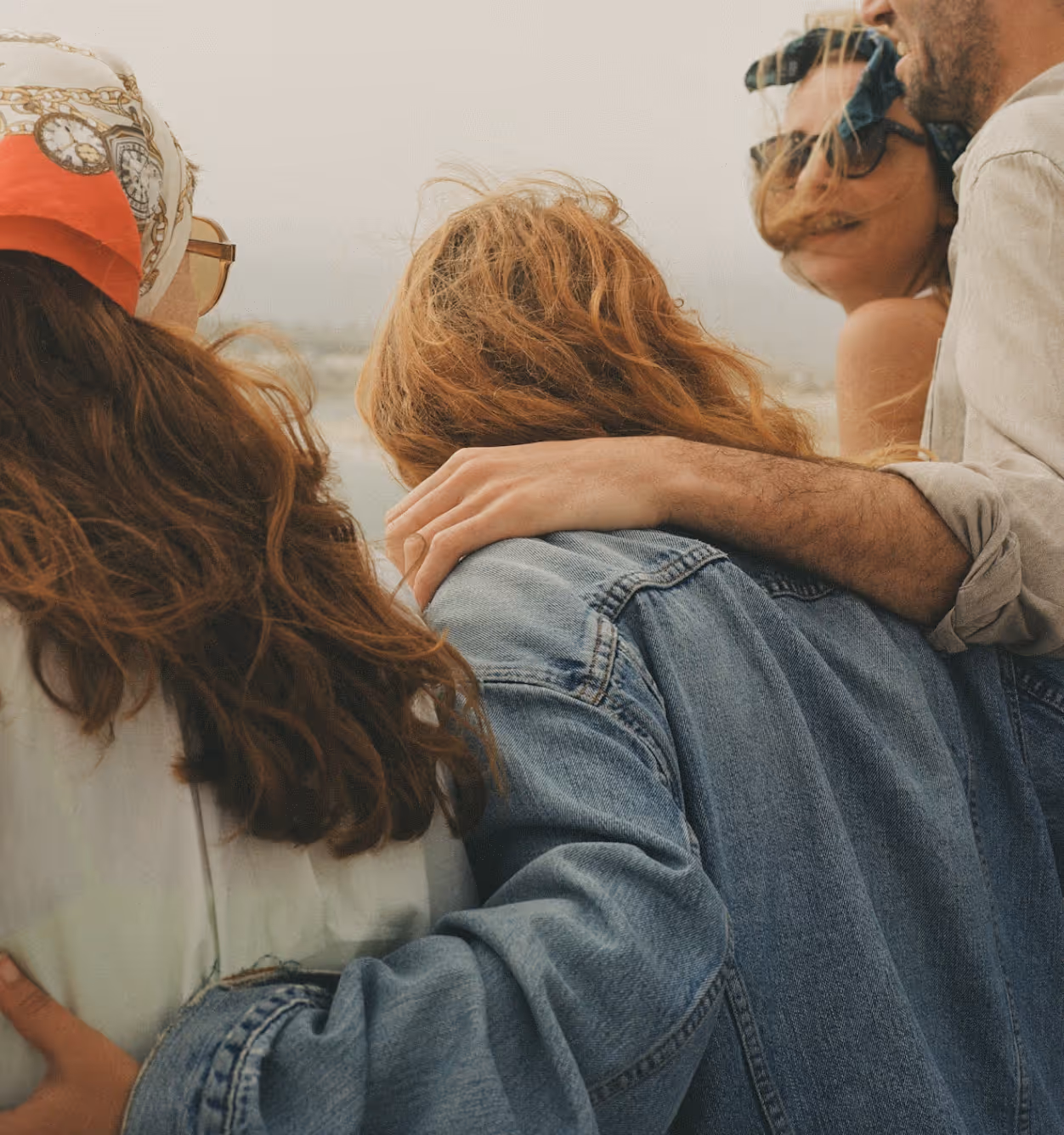 Four people stand close together outdoors, seen from behind and slightly to the side. Two have red hair and wear headscarves, one wears sunglasses, and one has an arm draped around another’s shoulders - all sharing a moment of connection and longevity.