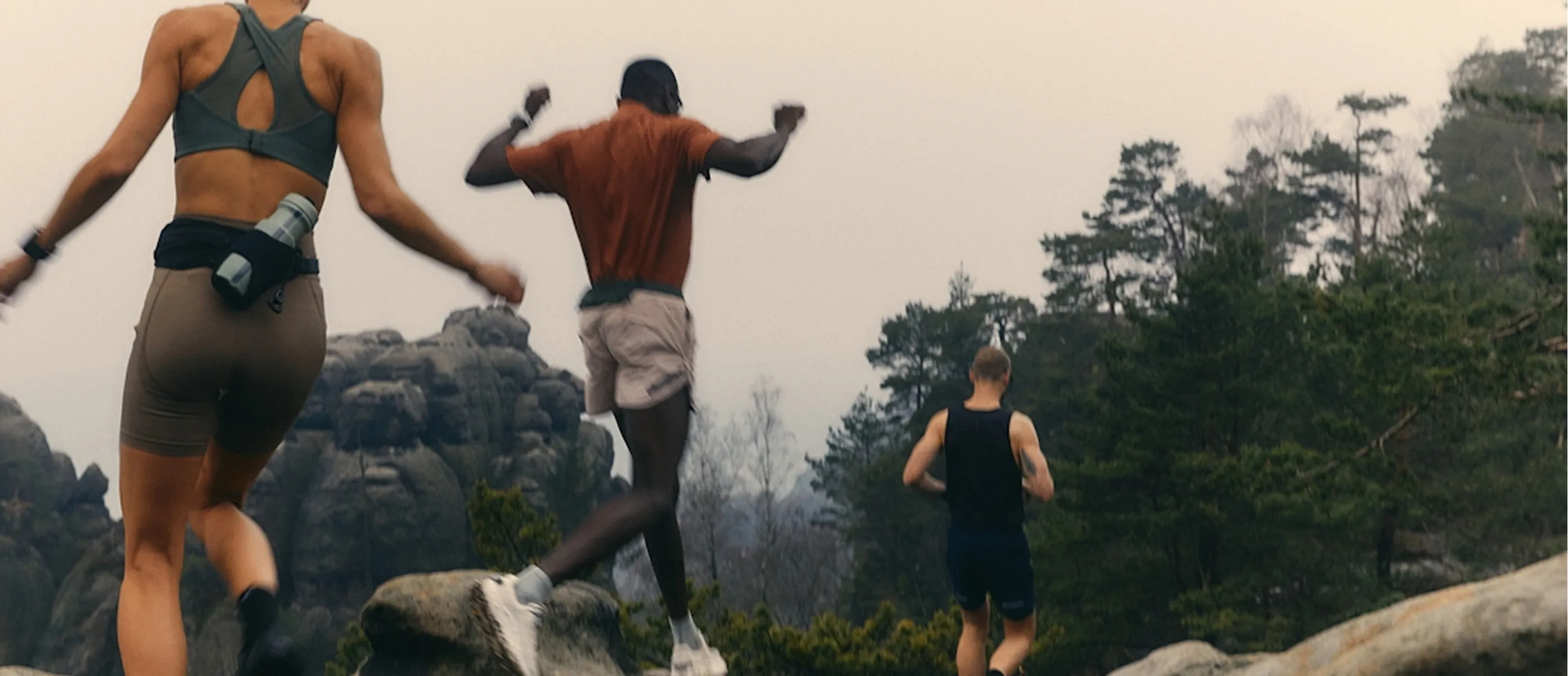 Three people in athletic wear run and jump over rocks in a forested, mountainous area under an overcast sky, highlighting the importance of outdoor activity for longevity.