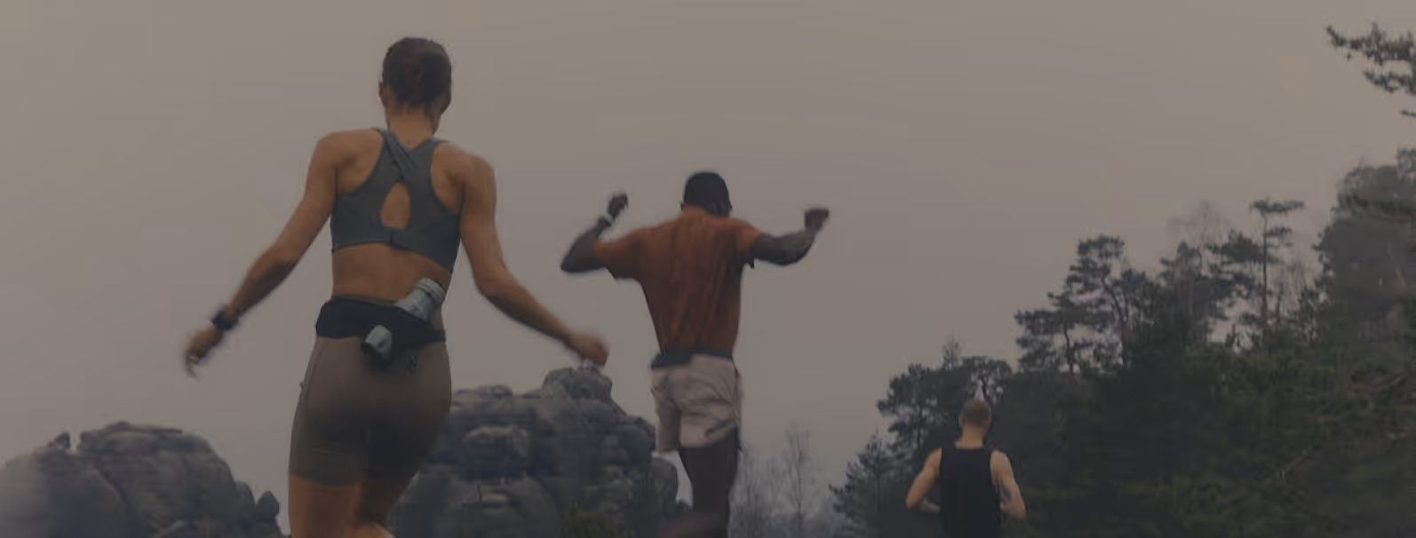 Three people running outdoors on a trail surrounded by trees and rock formations under a cloudy sky.