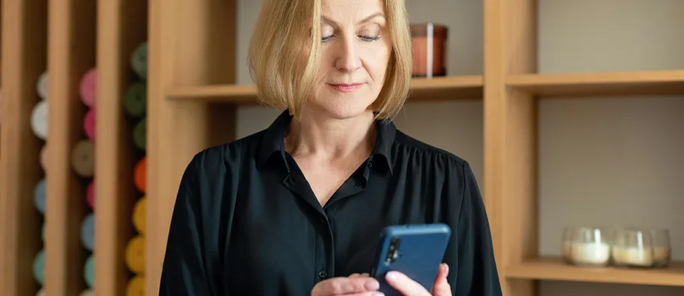 Mature woman in black shirt looking at blue smartphone in modern wooden shelving setting.