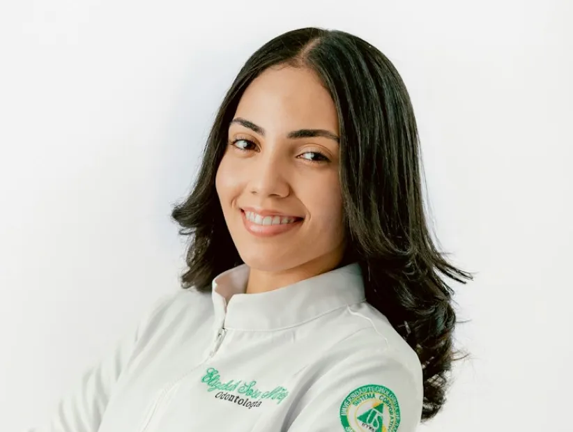 Smiling woman with long dark hair wearing white medical coat with green logo