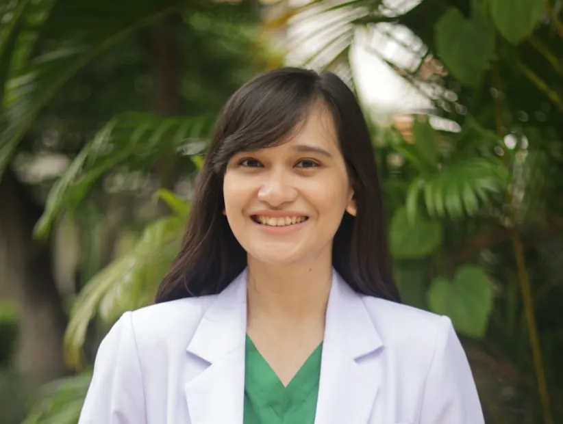 Smiling woman in white blazer and green top outdoors with foliage background
