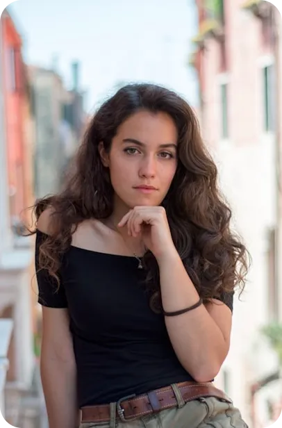Woman with long dark hair in black top poses thoughtfully in urban street setting.