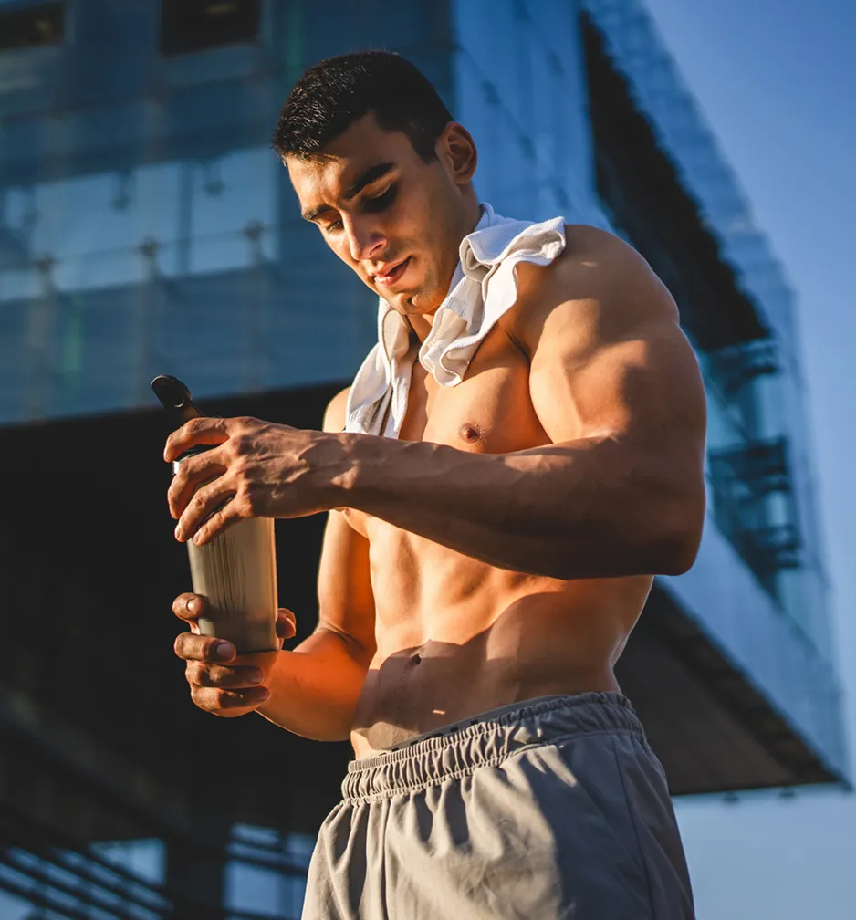 Muscular shirtless man outdoors with towel on shoulders opening a shaker bottle during workout.