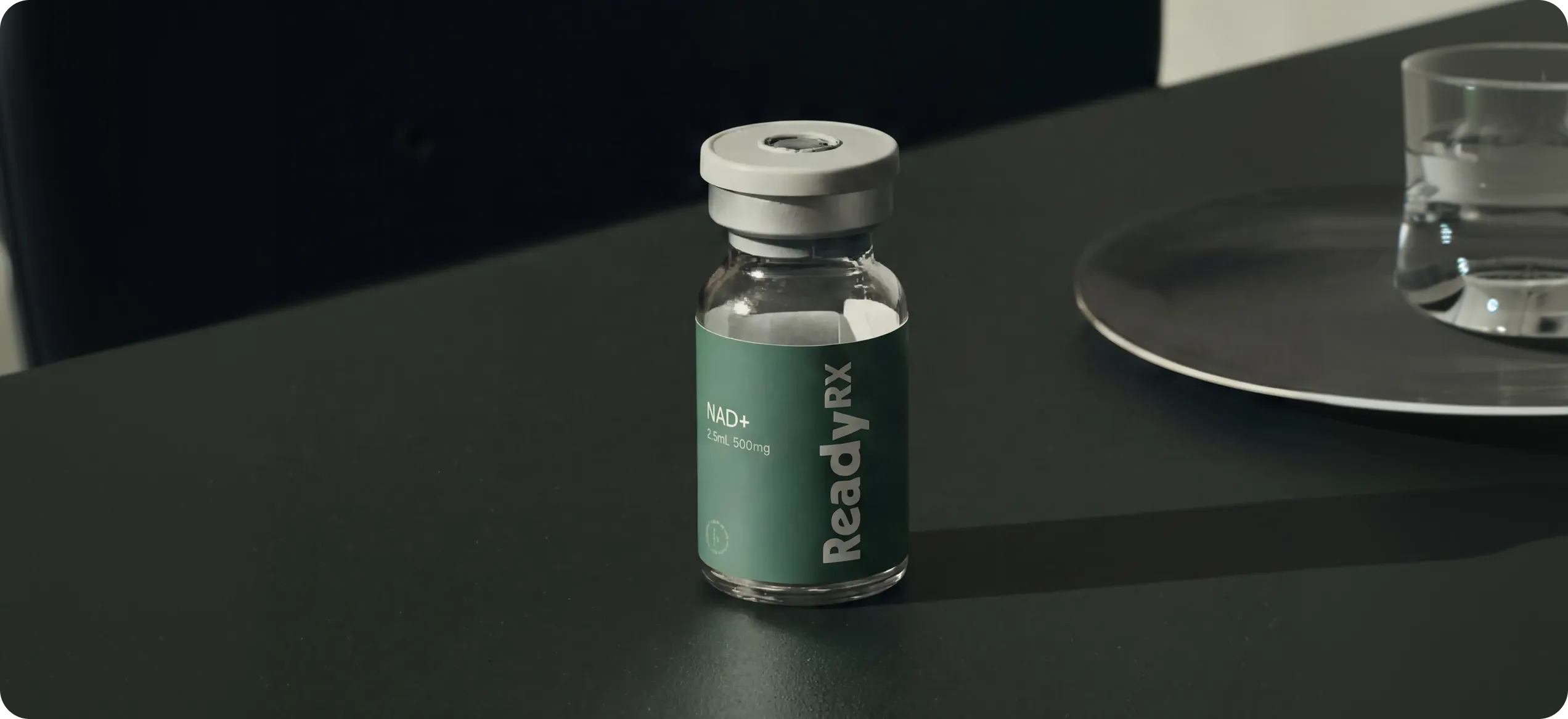 Clear vial labeled ReadyRX NAD+ 2.5mL 500mg on a dark surface with a glass of water nearby.