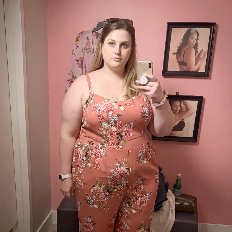 Woman in coral floral dress taking mirror selfie in pink bedroom with framed photos