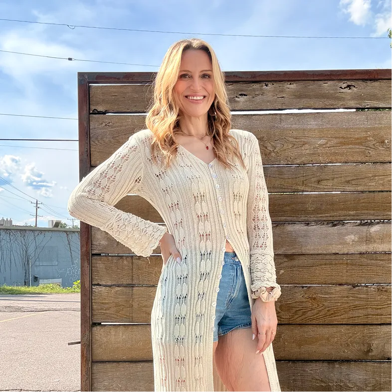 Woman smiling in beige cardigan and denim shorts against rustic wooden wall.