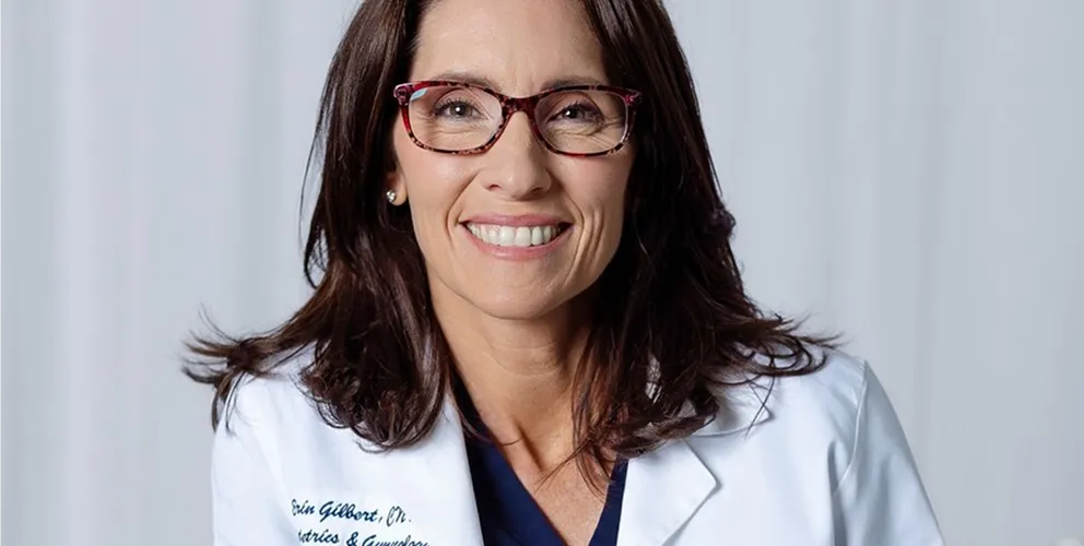 Woman with long dark hair wearing red-framed glasses and white medical coat, smiling