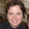 Smiling woman with dark hair wearing a dark colored top, looking at camera indoors