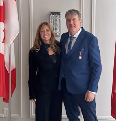 A smiling man in a blue suit with a medal and a woman in a black outfit standing together indoors with red and white flags in the background.