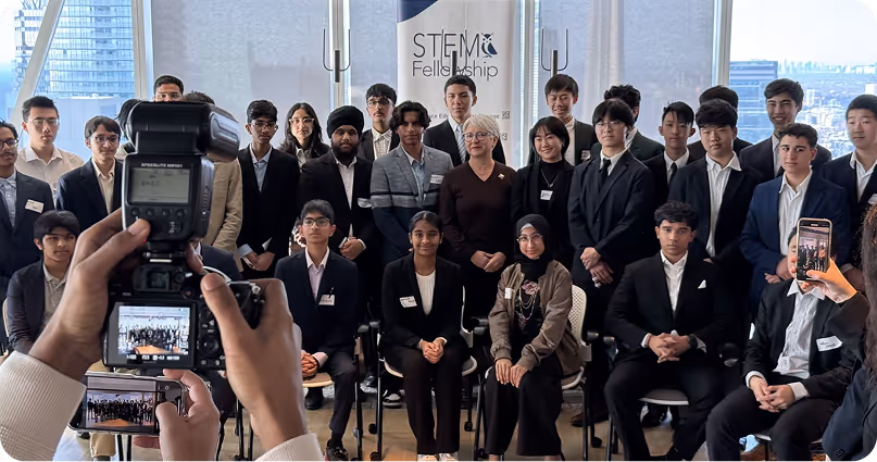 Group of diverse young adults and a few older individuals posing indoors for a STEM Fellowship group photo, with two people photographing them using a camera and a smartphone.