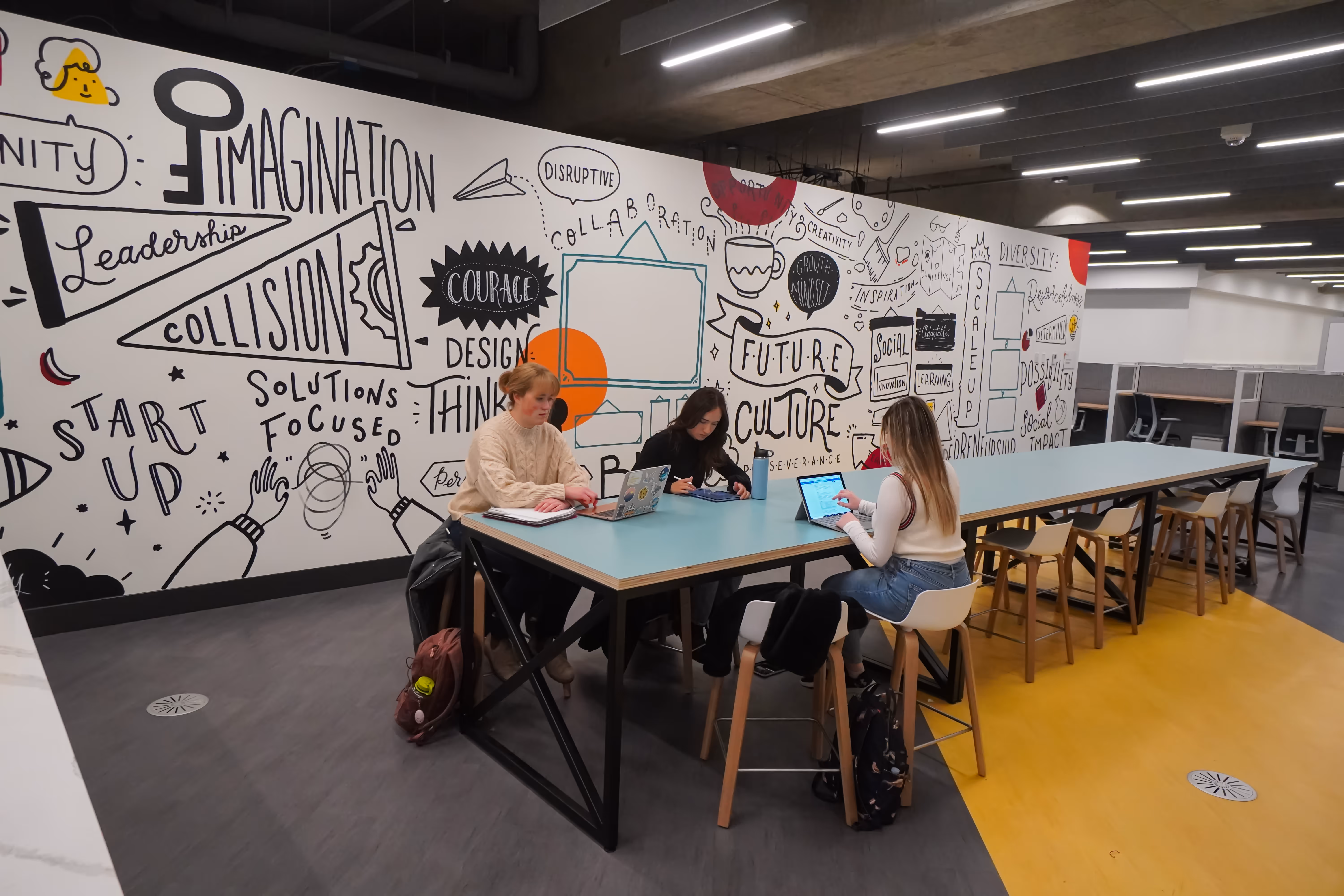 Three people working on laptops and tablets at a long table in a modern workspace with a mural featuring words like imagination, leadership, and culture on the wall behind them.