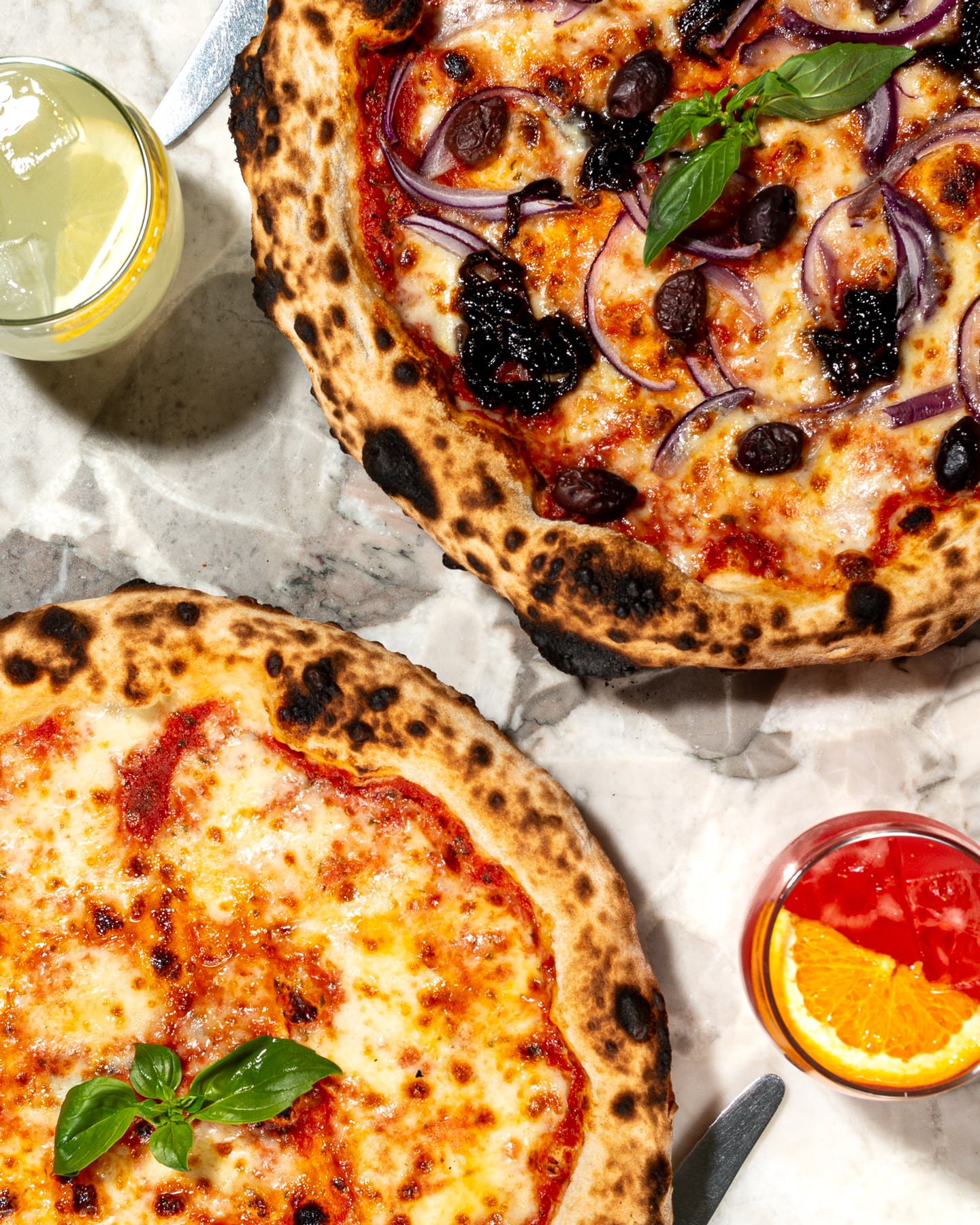 Authentic wood-fired pizzas at Paolo’s Italian Kitchen — a classic Margherita and a red onion with black olive pizza, paired with refreshing cocktails and lemonade on marble table.