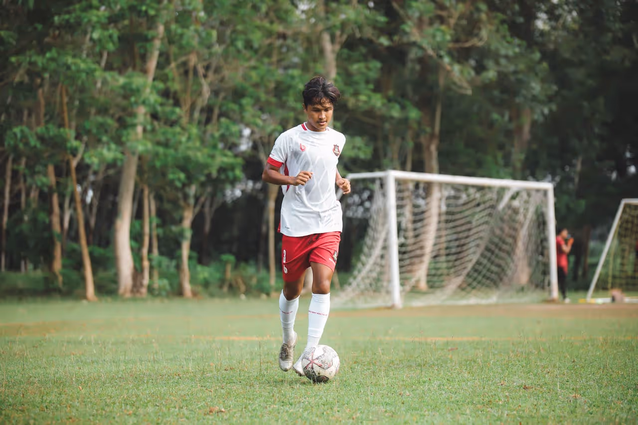 Young male soccer player wearing white and red uniform dribbling a soccer ball on a grassy field with goalposts and trees in the background.