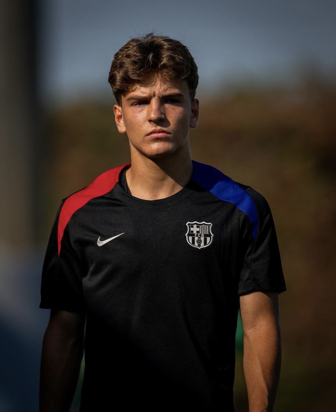 Young football player wearing a black Nike FC Barcelona training shirt with red and blue shoulder stripes, standing outdoors.
