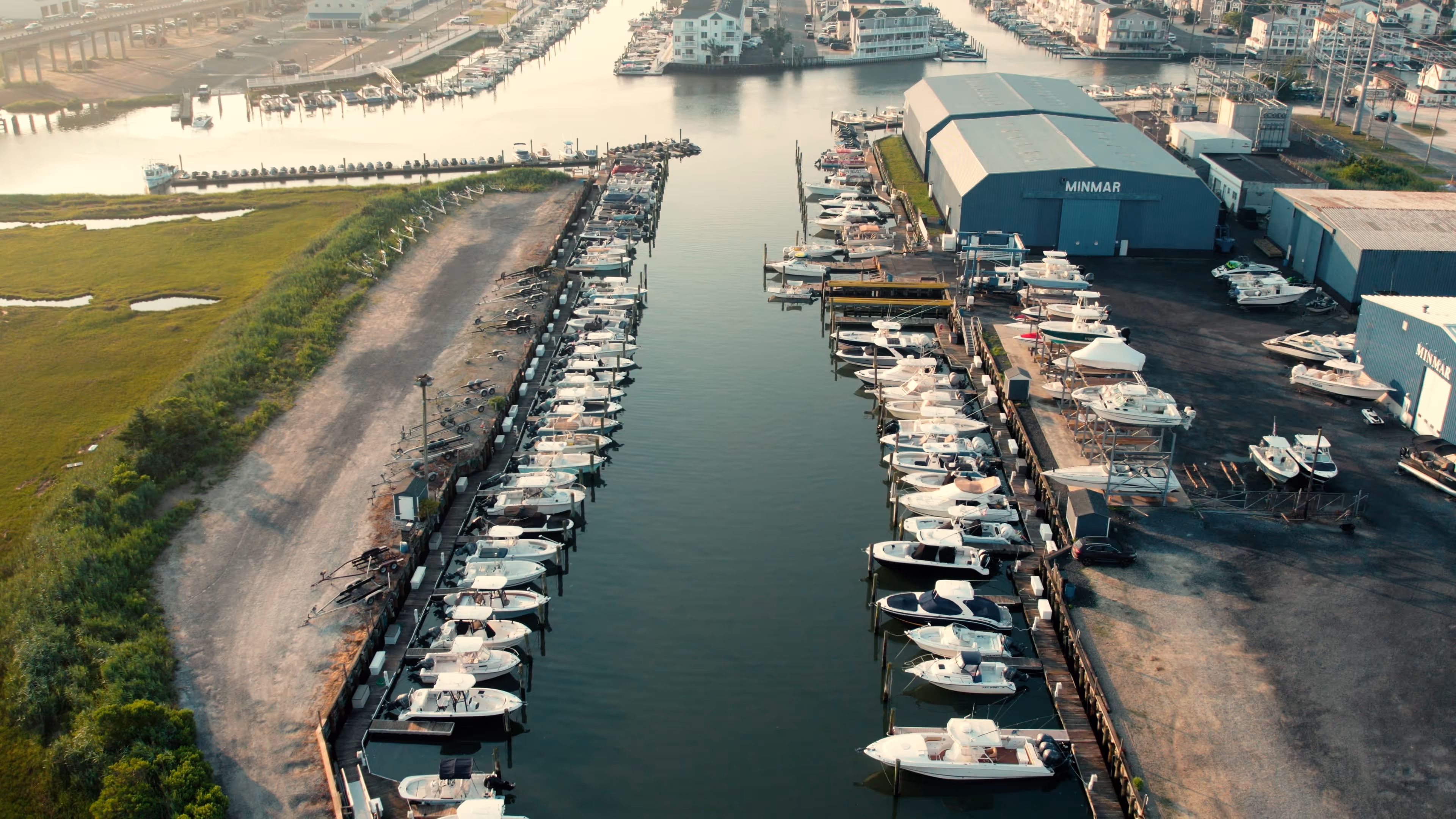 Aerial view of Minmar marina with many parked boats on both sides of a calm water channel, surrounded by green marshland and industrial buildings.