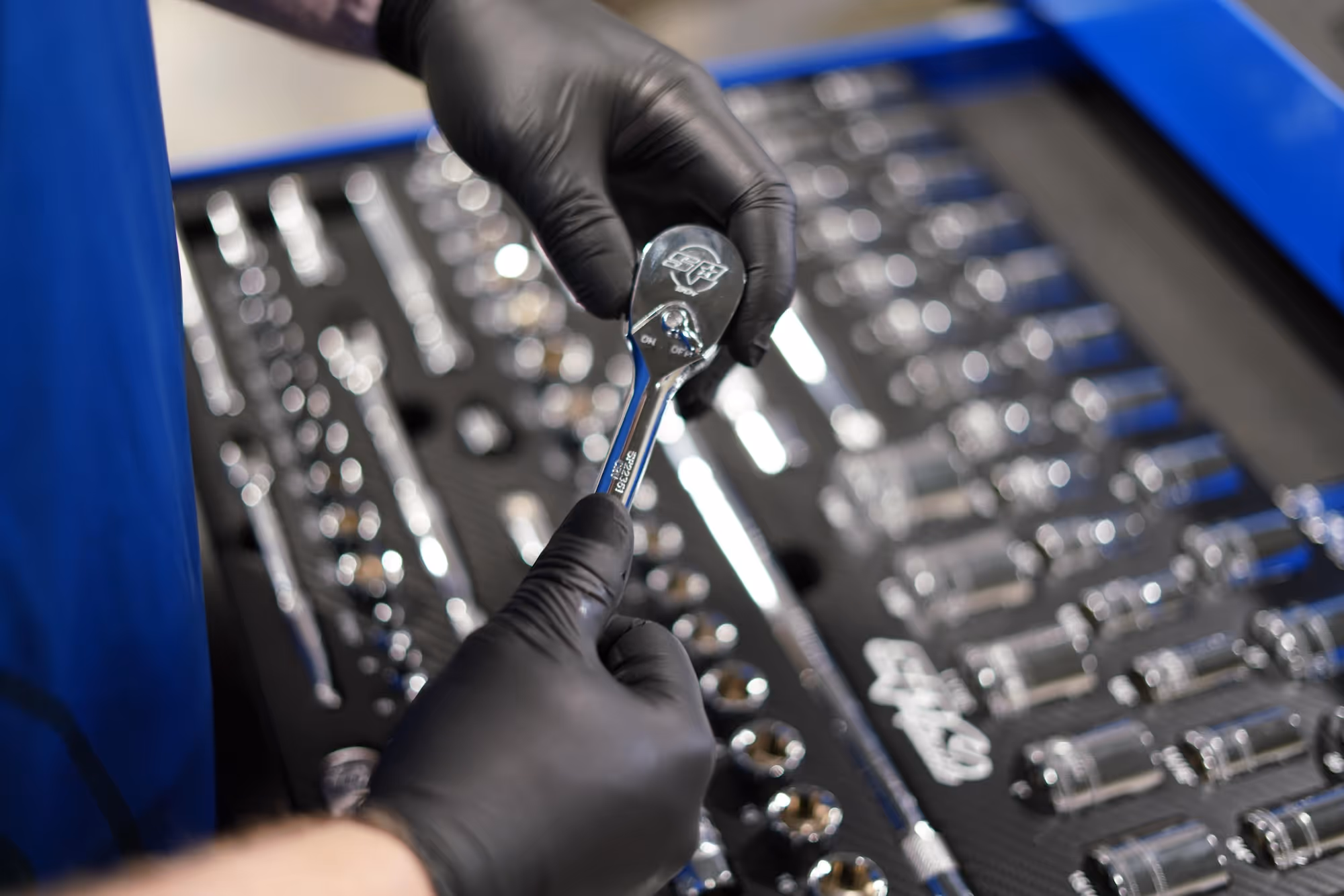 Person wearing black gloves holding a ratchet wrench above a toolbox with organized sockets and tools.