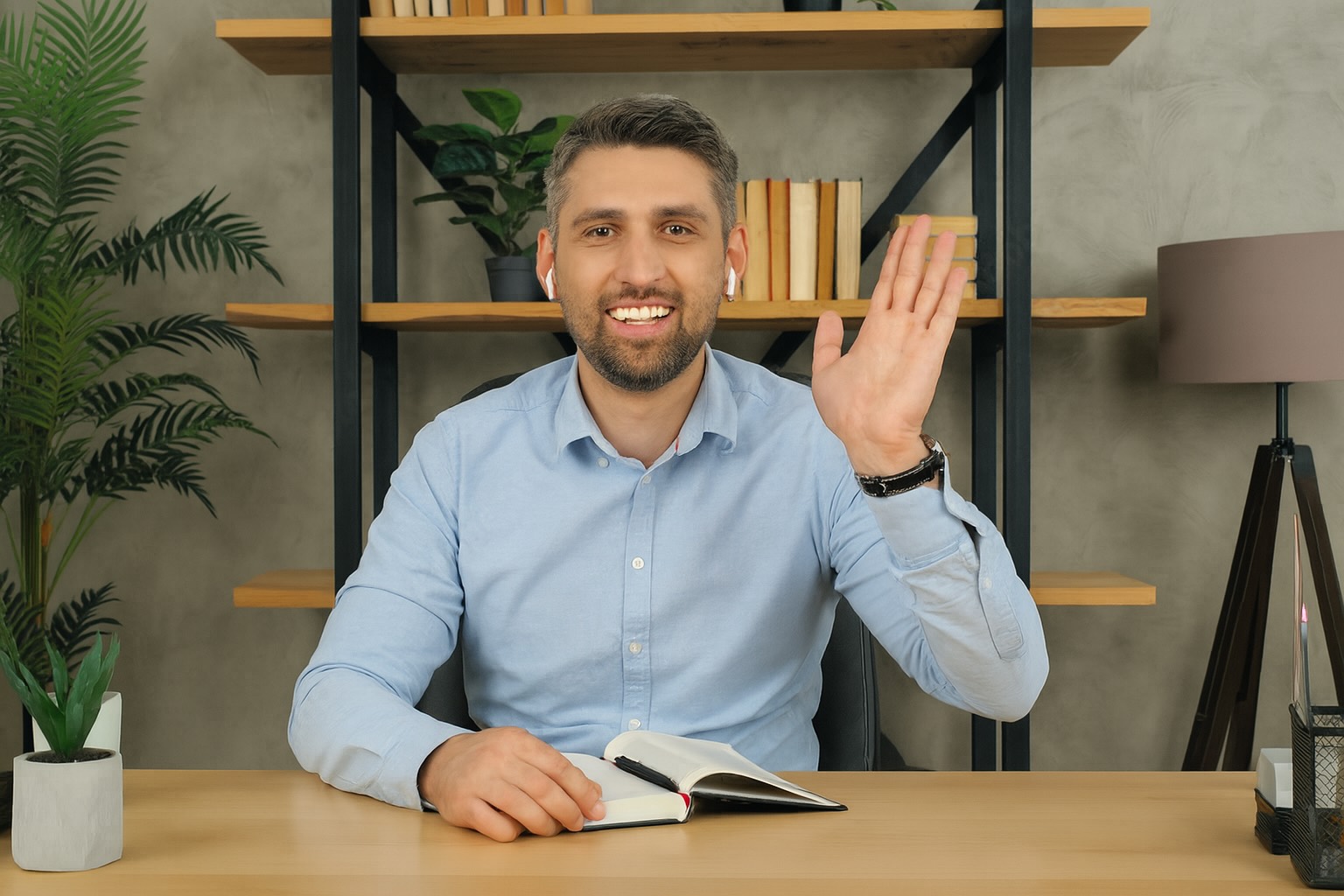 A smiling man in a light blue shirt sits at a desk, waving with one hand and holding an open notebook, with a bookshelf and plants in the background.
