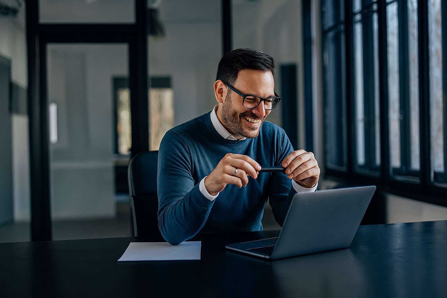 A smiling man in glasses and a teal sweater sits at a desk, holding a pen and looking at his laptop in a modern, sunlit office.