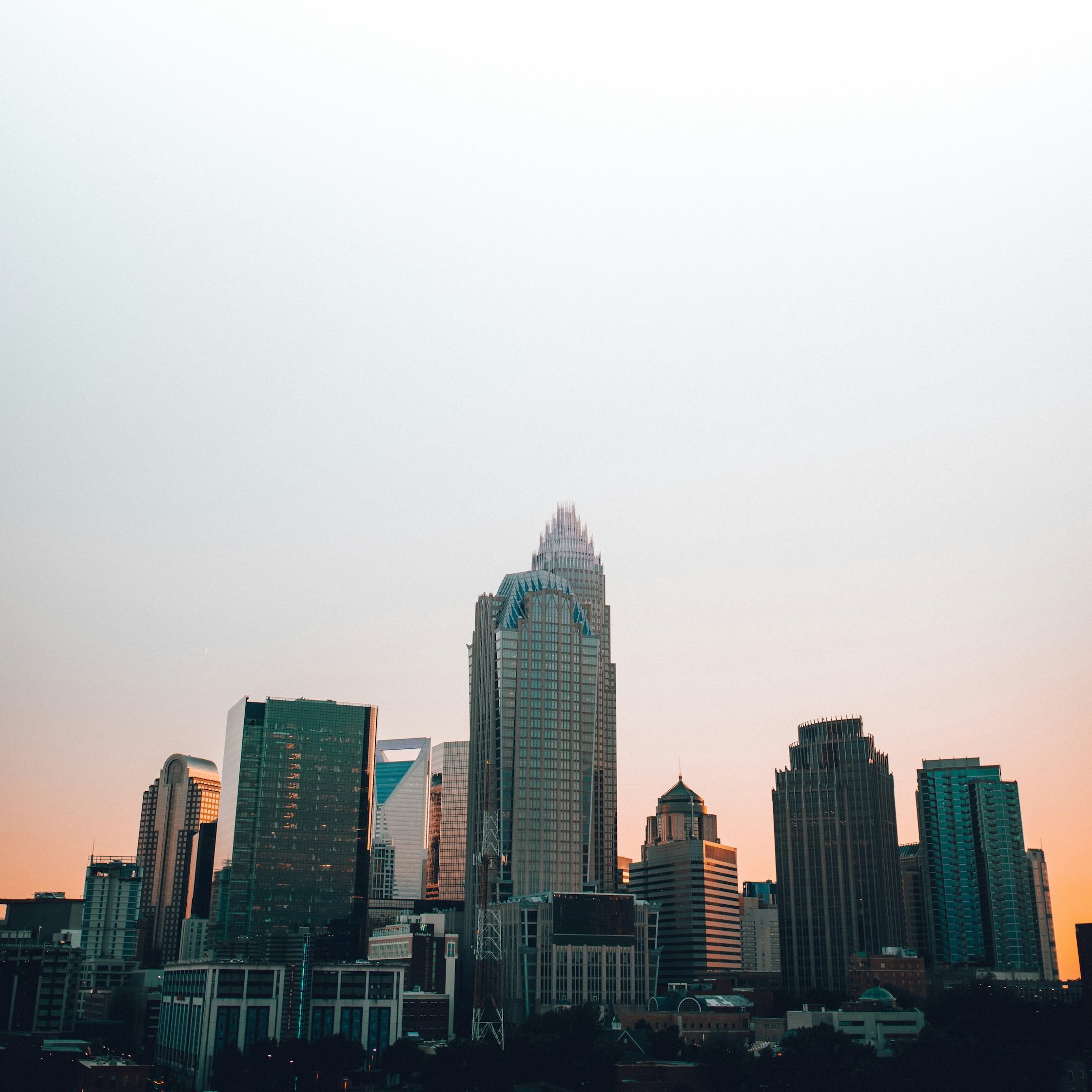 Downtown Charlotte skyline at sunset, featuring tall modern skyscrapers with a warm orange glow on the horizon.
