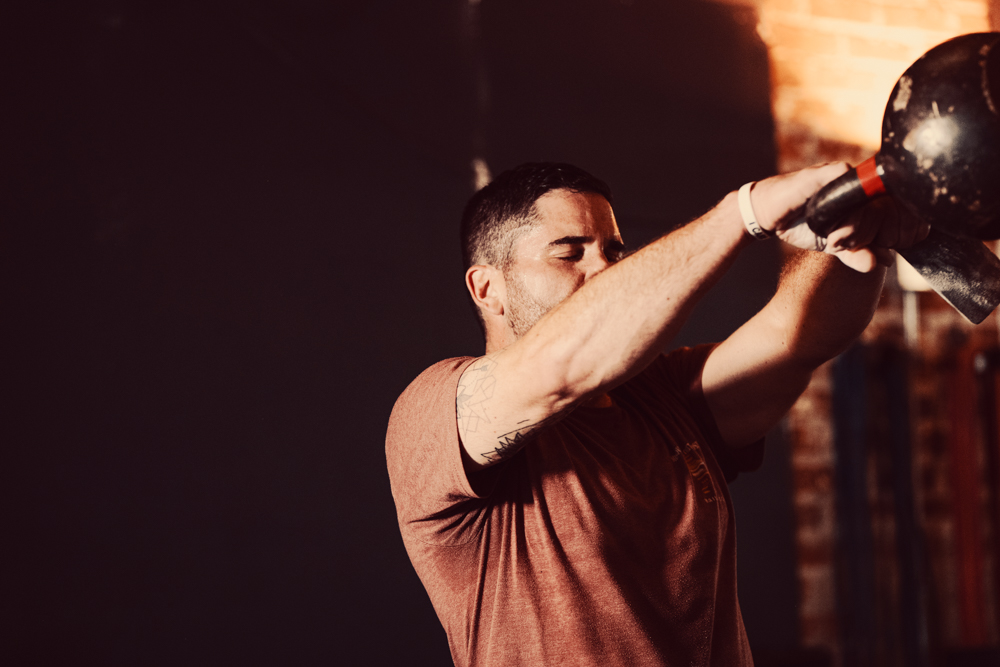 Man lifting a kettlebell in a gym with eyes closed, showing tattooed arm and wearing a brown shirt.
